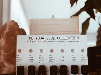 7 essential oil bottles in a row in front of a "yoga soul collection" chart which is leaned against a wooden box. A large crystal is in the background on the left.