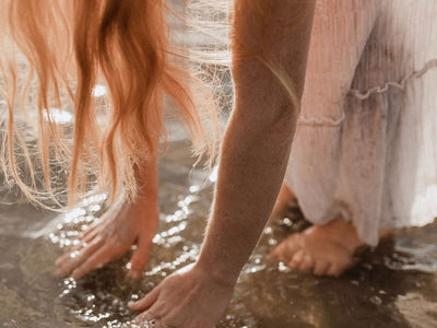 Woman in floaty dress standing in shallow sea water bending forwards with hands touching the sea and hair falling towards the water.